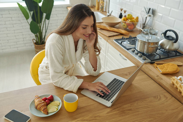 Top view of beautiful woman in bathrobe using laptop while having breakfast in the kitchen - Photo, image