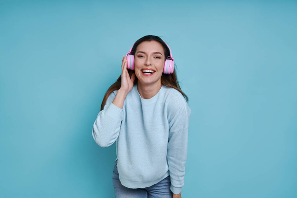 Beautiful woman in headphones listening music and smiling while standing against blue background - Fotografie, Obrázek