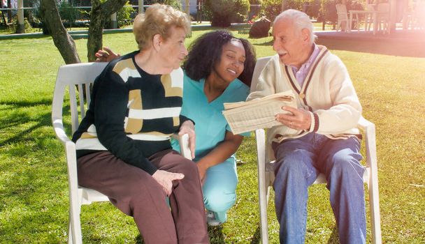 Afrikaanse verpleegster met behulp van tablet met bejaarde gepensioneerde echtpaar buiten in de revalidatie ziekenhuis tuin. Geluk, rehabilitatie en pensioen. - Foto, afbeelding