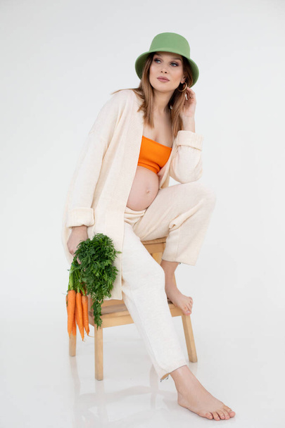 Γυναίκα με γυμνή κοιλιά στο τέλος της εγκυμοσύνης κάθονται στην καρέκλα κρατώντας δέσμη καρότο σε casual ρούχα με παναμά στο κεφάλι, λευκό φόντο. Πορτρέτο της νεαρής εγκύου περιμένει μωρό. Μητρότητα. - Φωτογραφία, εικόνα
