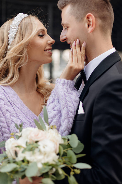 Beautiful young wedding couple in elegant outfits walk around city, embrace and kiss on street background. Portrait closeup of happy bride with bouquet and groom in love. Wedding day, family holiday. - Foto, Imagem