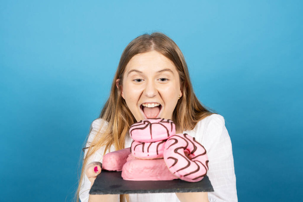 Mladá žena drží hromadu sladkostí sladké jídlo se závistí izolované na modrém pozadí.. Špatné stravovací návyky koncept. - Fotografie, Obrázek