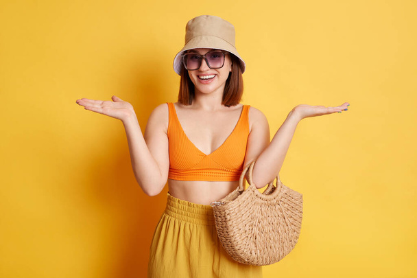 Horizontal shot of positive young young woman wearing swimsuit, shorts and panama, posing isolated over yellow background, standing spread arms, being not sure, don't know. - Zdjęcie, obraz