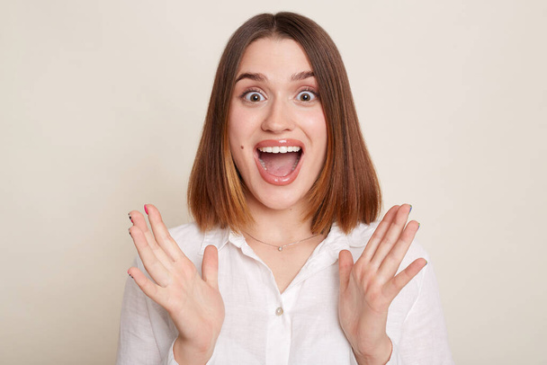 Horizontal shot of excited shocked woman wearing shirt posing isolated over white background, sees something amazed, raised arms, looking at camera with open mouth. - Photo, image