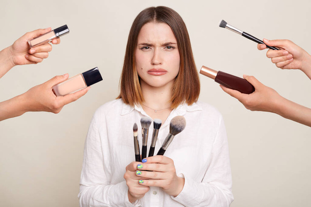 Image of the hands of several beauticians holding their respective equipment giving makeover to beautiful sad woman, standing with upset expression and sorrow, looking at camera with pout lips. - Foto, afbeelding