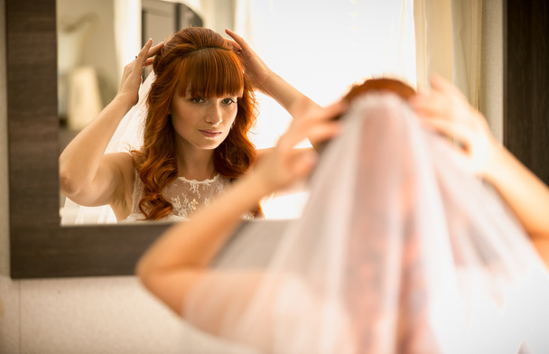 oblubienica piękny rudy patrząc w lustro i dołączanie welon - Zdjęcie, obraz