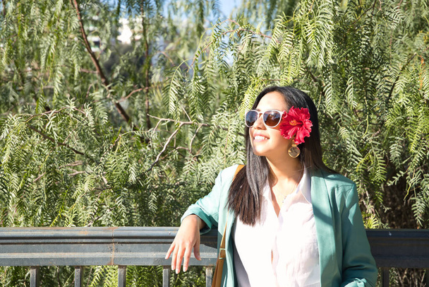 Fiatal és gyönyörű nő, dél-amerikai, zöld dzsekivel és napszemüveggel, piros virággal a fülében, zöld növényháttérrel. Koncepció utazás, szépség, virágok, természet. - Fotó, kép