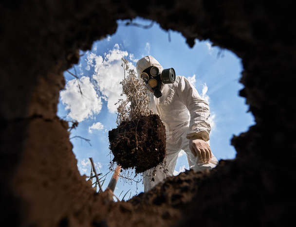 Vue de l'intérieur de la fosse de l'écologiste creusant la fosse par pelle et plantant un arbre, portant un masque à gaz et des combinaisons de protection à l'extérieur, sur fond de ciel nuageux bleu. - Photo, image