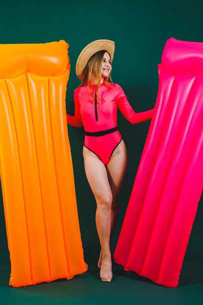 Happy pretty young woman in hat wearing bright pink swimsuit standing over isolated background holding inflatable pink mattress. Inflatable orange mattress. Summer vacation. - Photo, Image