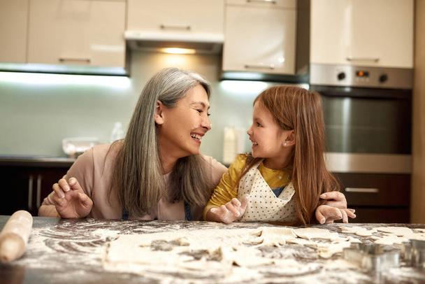 Grandmother and granddaughter with flour on faces looking at each other and laughing during cooking cookies. Concept of family education and enjoying time together. Homemade food cooking. Home kitchen - Foto, Imagen