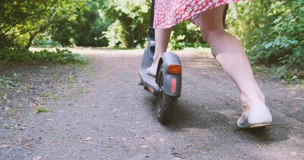 Donna in abito inizia a muoversi su scooter elettrico lungo il percorso nel parco. Si butta a terra con il piede. Vista posteriore, primavera estate, giorno, alberi verdi attraverso il fogliame il sole splende. Alto - Foto, immagini
