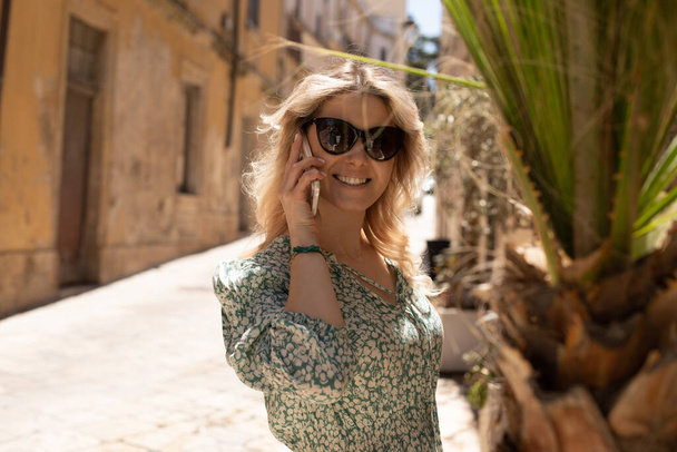 Portrait of young joyful beautiful woman with wavy fair hair wearing summer light green white dress, black sunglasses, standing on street near palm tree, old yellow buildings, talking on smartphone. - Photo, Image