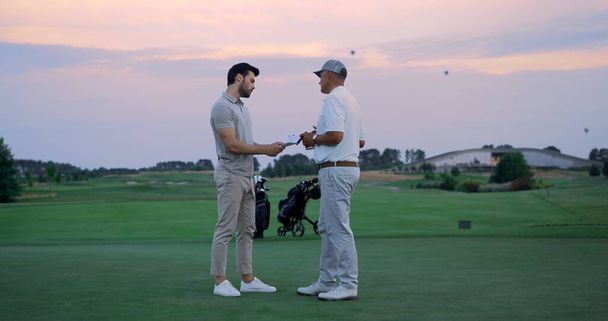 2人のビジネスマンがゴルフコースの外に立つ。スポーツマンはフィールドカントリークラブで論文に署名する。裕福なゴルファーチームは、日没の自然で一緒に合意を議論話をします.高級趣味パートナーのコンセプト - 写真・画像