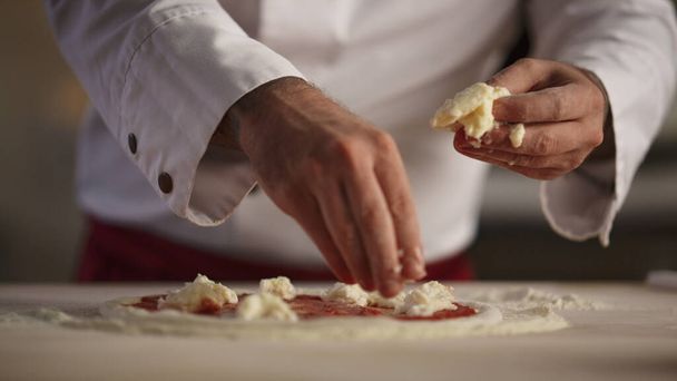 Pizza chef koken zelfgemaakte diner maaltijd in de keuken. Onherkenbare mannenhanden die mozzarella kaas in een professioneel restaurant van dichtbij leggen. Banketbakker toevoegen van ingrediënten. Concept van het voedselproductieproces. - Foto, afbeelding