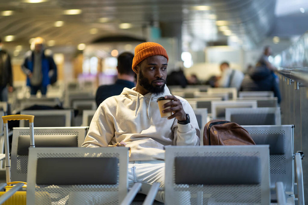Giovane viaggiatore afroamericano che beve caffè e mangia panino in attesa di volare al terminal dell'aeroporto. Trendy ragazzo hipster nero soddisfa la fame con un caffè e sandwich.  - Foto, immagini