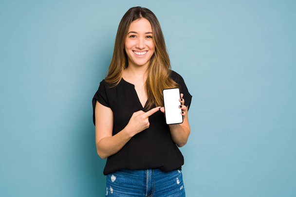 Mulher vestida casualmente apontando para uma tela branca em seu smartphone e sorrindo em um estúdio - Foto, Imagem