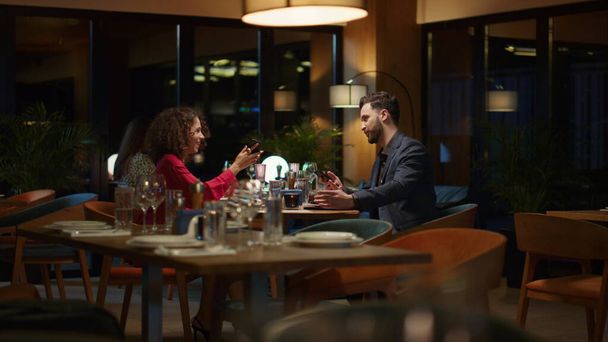 多民族のカップルは、レストランのテーブルで自撮り携帯電話を使用しています。2人の恋人は夜にモダンなスタイリッシュなカフェで記念日を祝う。彼女は携帯電話で写真を見せる。一緒に時間の概念. - 写真・画像