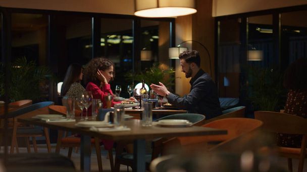 Πολυεθνικοί εραστές συζητούν μαζί σε ένα πολυτελές δείπνο σε εστιατόριο. Δύο ελκυστικοί άνθρωποι συνομιλούν στο φανταχτερό τραπέζι του καφέ. Οι εραστές απολαμβάνουν ο ένας τον άλλον στο μπαρ. Ρομαντικό δείπνο έννοια. - Φωτογραφία, εικόνα