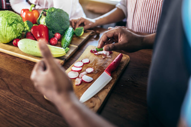Крупный план черного человека с острым ножом резки овощей на деревянной доске, делая свежий салат с использованием органических ингредиентов. - Фото, изображение