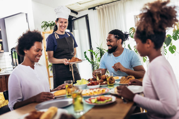 Une famille afro-américaine multigénérationnelle savourant à table. Femme blanche cuisinier les servir. - Photo, image