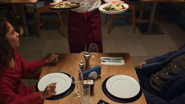 Waiter serving restaurant food to multiethnic couple at romantic dinner date. Kitchen chef putting plates on table close up. Love partners going out in fancy cafe bar. Elegant dating enjoyment concept - Photo, Image