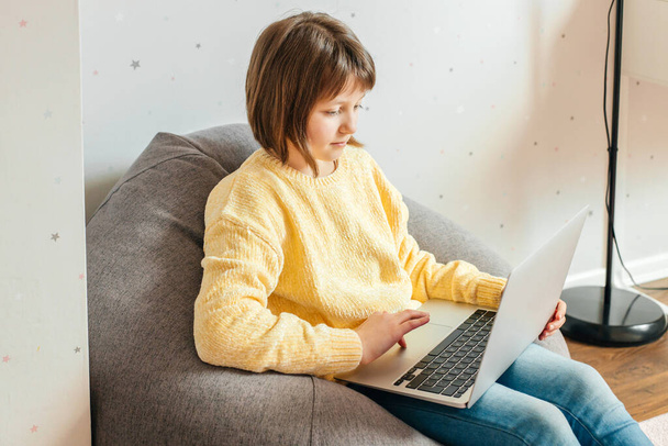 Ein Mädchen sitzt mit einem Laptop auf dem Schoß in ihrem Zimmer und schaut Unterricht oder Schulungen. Kinder nutzen Technologie. Online lernen und lernen - Foto, Bild