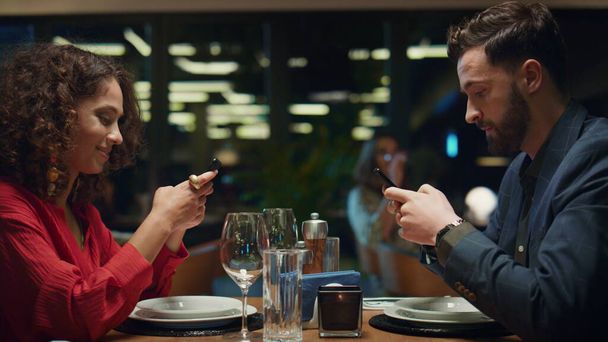 Multiethnic couple texting mobile phone in fancy restaurant table. Two mixed race couple browsing internet in hotel bar. Love partners surfing social media in luxury cafe place. Dinner date concept. - Fotó, kép