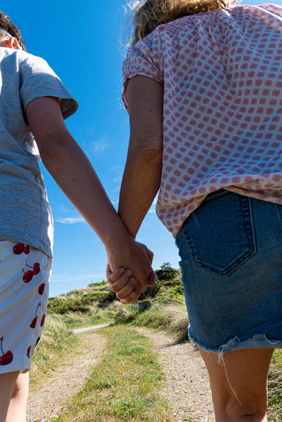 Hirtshals, Denemarken Een moeder en zoon lopen hand in hand een onverharde weg naar het strand met handdoeken . - Foto, afbeelding