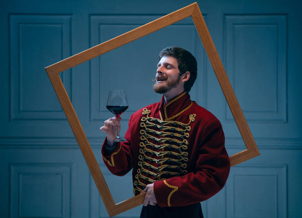 Astonished warrior tasting red wine. Creative portrait of young man in image of medieval hussar posing with picture frame isolated on dark blue background. Retro style, comparison of eras concept. - Foto, afbeelding