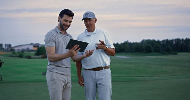 Dos golfistas buscan tableta en el paisaje de campo. Hombres felices charlando en línea afuera. Jugadores de golf alegres sonriendo en divertido fin de semana de actividad deportiva. Empresarios hablando del dispositivo de la almohadilla. Concepto de lujo. - Foto, Imagen