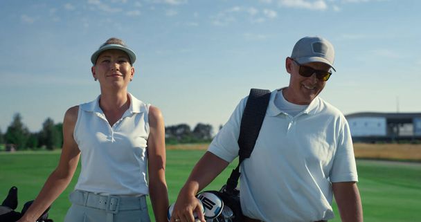 Dışarıda golf oynayan zengin bir çift. İki golf kulübü üyesi sahada yürüyor. Neşeli golfçüler güneşli yaz gününde sohbet ediyorlar. Kapatıyorlar. Rahat koç takımı maçta golf sopaları taşıyor. Hobi konsepti. - Fotoğraf, Görsel