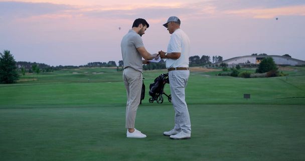 Boze golfspelers tekenen een contract op de green course. Sportteam ruziën op country club. Twee golfers praten over buiten scoren. Geïrriteerde sporters gooien dossiers over de natuur. Probleemconcept. - Foto, afbeelding