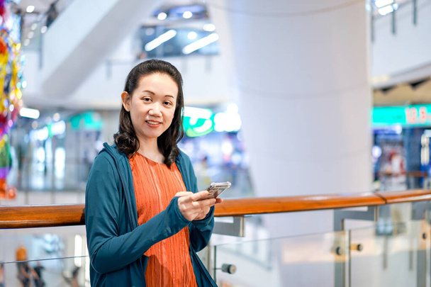 Usmívající se asijská žena Podívá se na fotoaparát s držení smartphone v ruce, Stojí v oddělení Store Atmosféra. - Fotografie, Obrázek