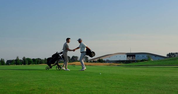 Golfspelers ontmoeten elkaar op het terrein. Golfteam praat buiten over sport. Twee golfers schudden elkaar de hand op het platteland. Vrienden genieten van weekend activiteiten. Vrijetijdsconcept. - Foto, afbeelding