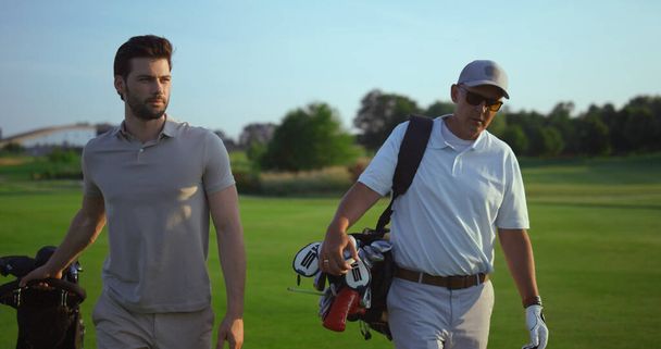 Handsome golf players talk sport hobby. Active men carry golfing putters clubs outdoors. Successful professional golfers walking on green grass club field on summer weekend. Sportsmen together concept - Photo, Image
