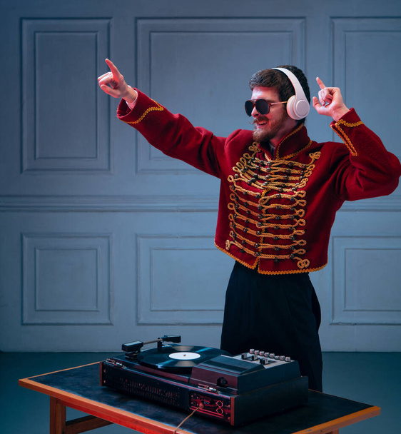 Eras comparison. Excited young man, dj wearing retro hussar costume playing, making sounds isolated over blue backgroundght. Nightclub performance. Concept of party, music, youth culture, ad - Foto, imagen