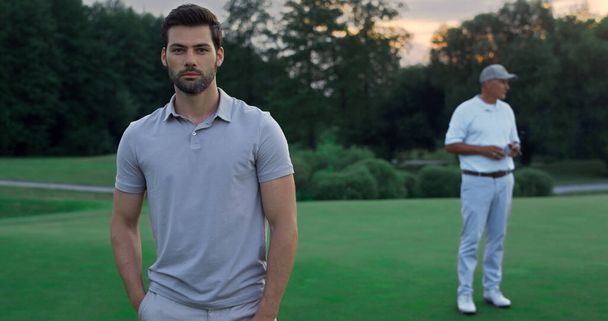 Focused model enjoy golf on sport team outdoors. Two players standing on green course. Attractive bearded guy posing at sunset fairway. Group men rest at luxury country club. Relax golfing concept. - Photo, Image