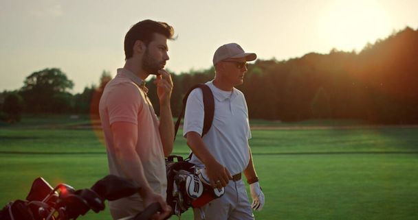 Πλούσιοι που παίζουν γκολφ στο ηλιοβασίλεμα. Δύο παίκτες απολαμβάνουν τον αθλητισμό στο γήπεδο. Επαγγελματική ομάδα παίκτες αναζητούν απόσταση στην ύπαιθρο club. Ενεργό ομαδικό σταντ στα αθλητικά. Έννοια αναψυχής. - Φωτογραφία, εικόνα