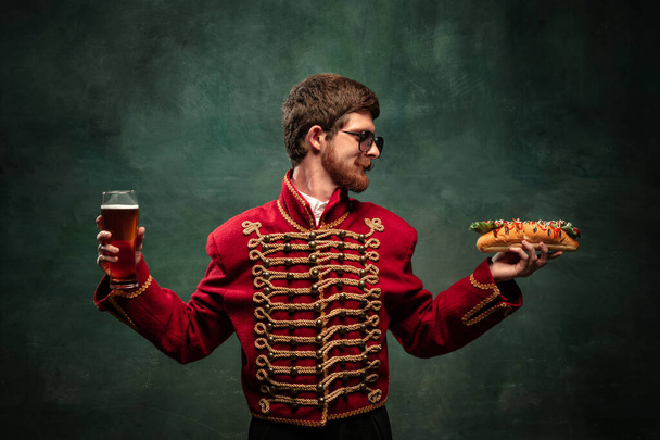 Young man in suit as royal hussar tasting big hot-dog isolated on dark green background. Handsome male model like historical character, old-fashioned hero. Concept of art, beauty, creativity, humor - Фото, изображение