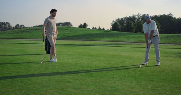 Golfing team play sport on course. Golf group practicing hits in country club. Professional players swing clubs putter hitting ball outdoors. Two golfers training skills on fairway. Luxury concept. - 写真・画像