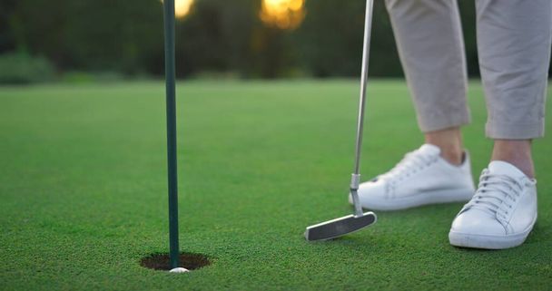 Die Beine des Golfspielers entspannen sich am Golfball-Loch. Golfer posiert auf grünem Platz aus nächster Nähe. Der Unbekannte trug Sportschuhe. Sportler genießen erfolgreiche Birdie Score auf dem Fairway. Golf-Luxus-Konzept. - Foto, Bild