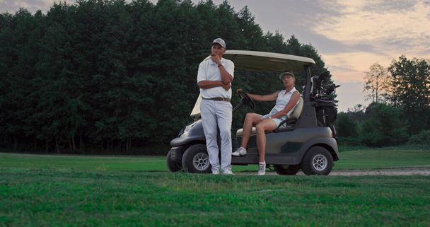 2人のゴルファーはゴルフコースで屋外で休む。夫婦でゴルフカートでリラックス。フェアウェイゲームでルックカメラをポーズ裕福なスポーツチーム。プロの選手は夏の夕日を楽しむ。週末のコンセプト. - 写真・画像