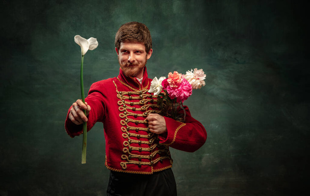Cavalier and beau. Handsome young man in image of medieval hussar holding flowers isolated on dark blue background. Retro style, comparison of eras concept. Male model wearing hussar uniform. - 写真・画像