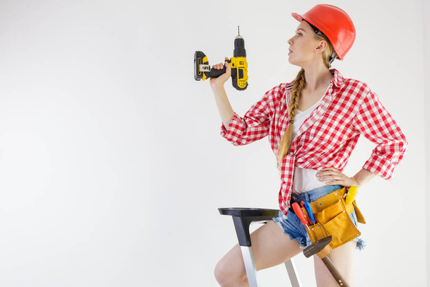 Young woman on ladder with toolbelt using driil and some power tools for her work at home. Girl working at flat remodeling. Building and renovation. - Foto, immagini