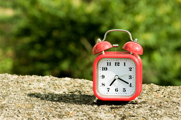 Reloj despertador rojo de estilo retro en una superficie de hormigón con árboles borrosos en el fondo. - Foto, imagen