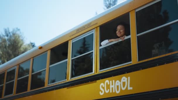 Uśmiechnięta Afroamerykanka wyglądająca rano przez okno szkolnego autobusu. Kręcone słodkie uczennica wieku podstawowego patrząc w oddali sam. Szczęśliwy uczennica stojący żółty pojazd autobus szkoły pojazdu.  - Materiał filmowy, wideo