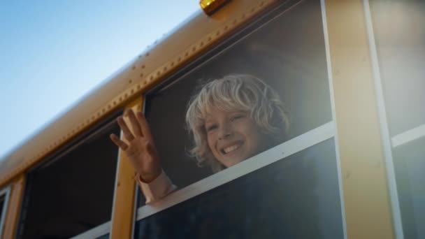 Mosolygó iskolás gyerek integet ki az iskolabusz ablakon egyedül közelről. Vidám, göndör fiú elbúcsúzik anyától. Vidám általános iskolás diákok elmennek nyári órákra. Boldog gyermekkori koncepció.  - Felvétel, videó