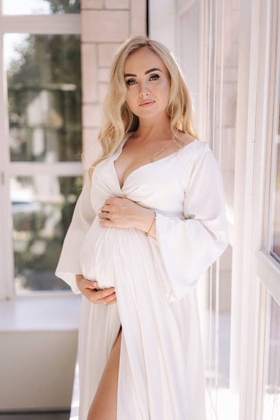 Beautiful portrait of pregnant woman in white dress. Concept of perfect pregnancy. Woman put her hand on belly. Future mother is waiting for baby. - Photo, Image
