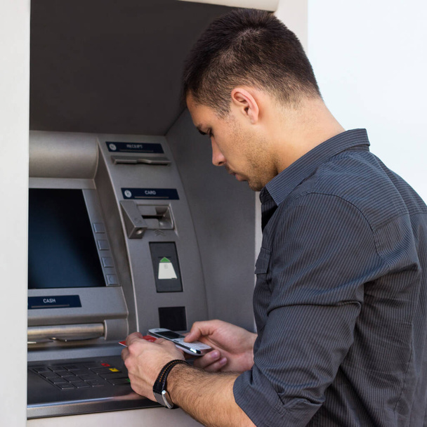 Junger Mann telefoniert und legt seine Kreditkarte am Geldautomaten ab - Foto, Bild