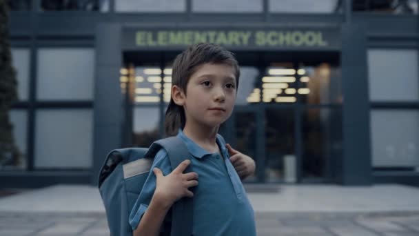 Aranyos tini fiú áll a modern iskola bejáratánál és megjavítja a hatalmas hátizsákot közelről. Egy általános iskolás fiú portréja, aki nehéz táskával pózol. Komoly kis fickó készen áll a tanulásra.. - Felvétel, videó